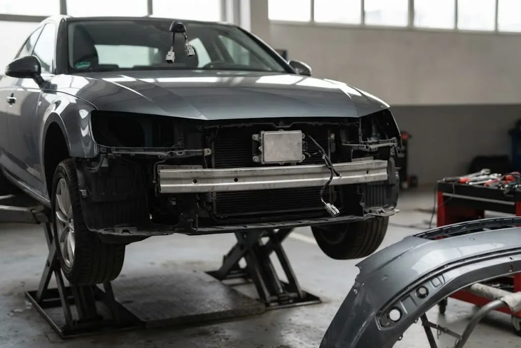 A vehicle in a calibration bay with its front bumper removed, exposing the long-range radar module behind the grille and a camera bracket at the top of the windscreen, with the ultrasonic sensor apertures visible in the removed bumper resting on a stand alongside and a patterned calibration target on a frame in front of the car.