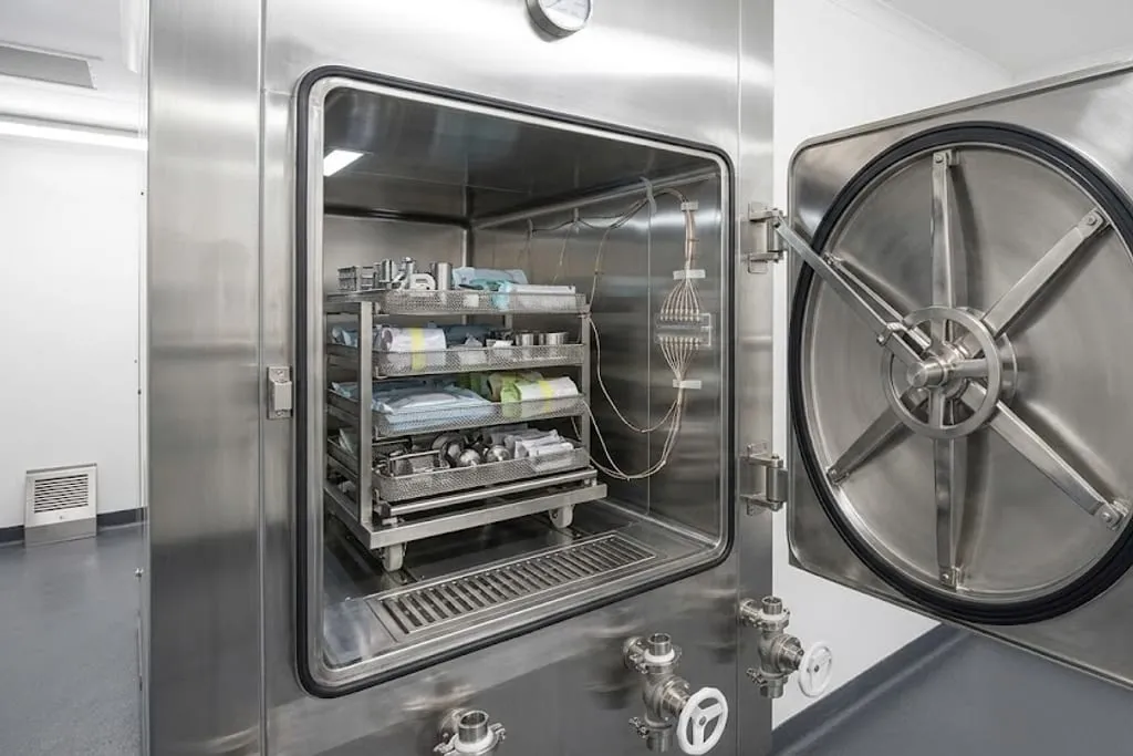 A large autoclave with its chamber door swung open, the heavy radial locking wheel on the inner face of the door. Wrapped packs and perforated stainless trays sit on a loading trolley inside the stainless chamber, and a bundle of fine thermocouple wires runs from the load out through a port in the chamber wall. A grille covers the drain in the chamber floor.
