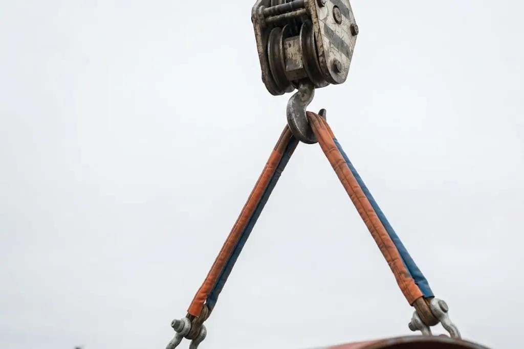 A close view looking up at a crane hook block with two round sling legs running down from the hook at a wide spread angle to shackles at the top of a large pipe, the legs clearly far from vertical against a bright overcast sky.