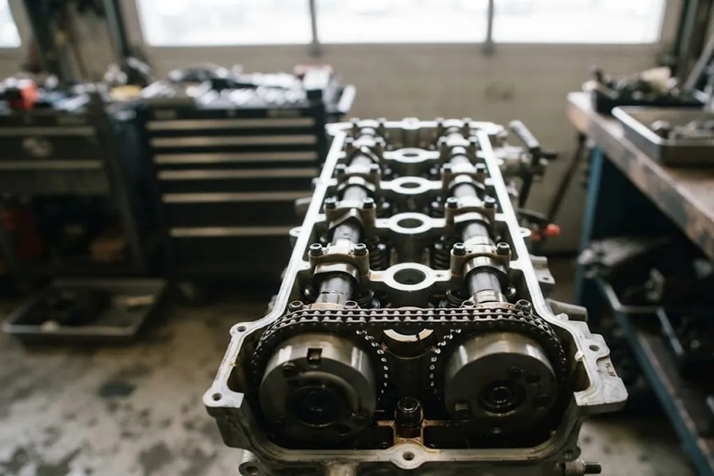 A close view of a twin camshaft cylinder head with the cam cover removed, showing both camshafts, the cam lobes bearing on bucket tappets over the valve springs, and the timing chain sprockets with a hydraulic cam phaser unit on the inlet camshaft.